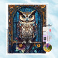 5D Diamond Painting Motiv: Eine majestätische Eule sitzt auf einem Ast mit Blumen, wunderschön umrahmt von einem klassischen blauen Buntglas-Fenster mit goldenen Ornamenten.